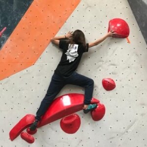 Un grimpeur escalade un mur d'escalade intérieur, s'agrippant à des prises rouges tout en portant un t-shirt noir Adrénaline et un pantalon foncé.