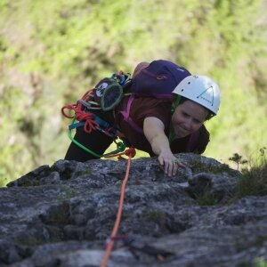 Une grimpeuse atteignant une prise tout en ascendant une paroi rocheuse verticale, portant un casque et attaché à une corde.